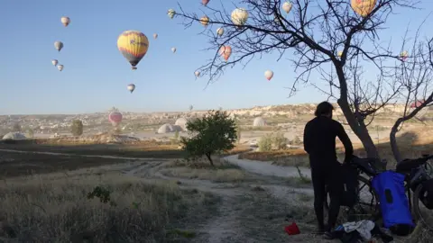 Sam Murray A man standing by a tree and he is looking at hot air balloons across a field.