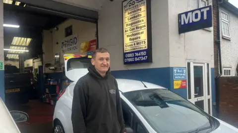 BBC/ADRIAN HARMS Shane Fry stands in front of a white hatchback car in the entrance to his garage in Godstone. He wears a dark branded fleece with DD Motors embossed on it.