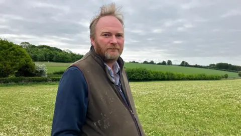 George Brown stands looking towards the camera, wearing a brown fleece gilet, a dark blue jumper underneath and a blue collared shirt. He has ginger hair and a beard. Behind him are green fields, hills and trees.