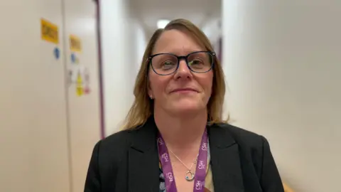 Chief executive officer, Renée Padfield, looks straight at the camera. She is wearing a dark suit jacket and purple lanyard and standing in a corridor with beige walls.