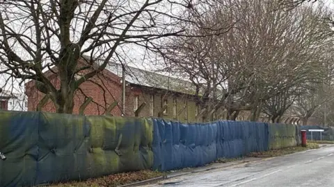 Simon Jones/BBC A tall fence with blue tarp running along it. There is a brick building and trees the other side of the fence.