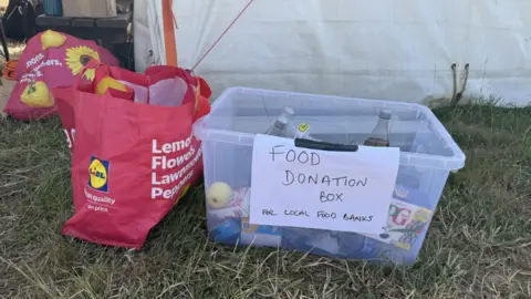 A clear plastic box with a handwritten paper sign attached to it saying "Food donation box for local food banks" on the grass outside a tent in a campsite at Glastonbury. It contains some bottles of soft drink, some tinned food and a box of tea bags. There is a red Lidl Bag for Life next to it.