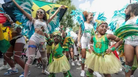 PA Media Performers in colourful carnival costumes, featuring green, blue, white and gold feathers and sequined details, take part in a street parade, with children and adults dancing together.