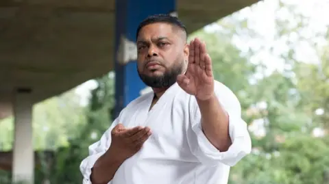 Shelim Ali outside under a flyover. He is doing a karate pose, holding both his hands up. He is looking directly at the camera.