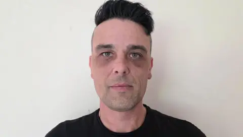 Kevin Brewer has black hair. He's wearing a black top and standing against a white wall.