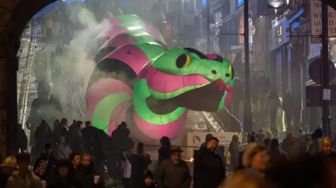 Shows a large illuminated snake in green, pink and black going down Shipquay Street, with people in the foreground in front of an arch.