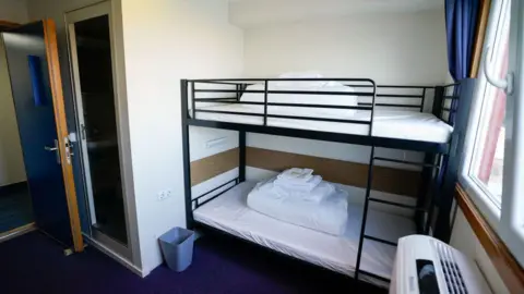 Getty Images A basic room with a bunk bed and clean sheets in a pile on top of the beds. There is a radiator on the wall and a bin by the bed frame. There is a window on the wall