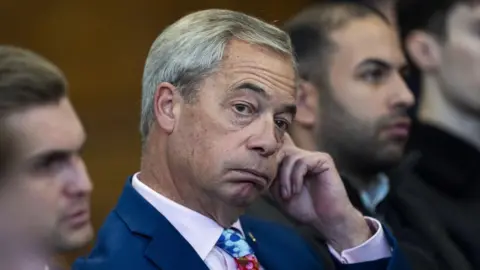 PA Media Nigel Farage wears a bright blue suit, pink shirt and blue patterned tie, looking off camera. He is puffing his cheeks.