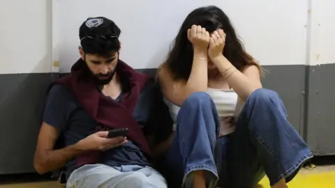 A man looks at his phone and a woman next to him covers her face with her hands in a bomb shelter in Tel Aviv (18/03/26)