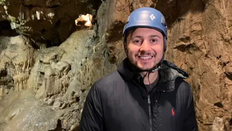 A man in a caving helmet stands in front of a collection of stalagmites and stalactites
