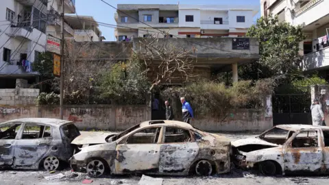 As pessoas da Reuters passam por carros queimados em Jableh, oeste da Síria (12 de março de 2025)