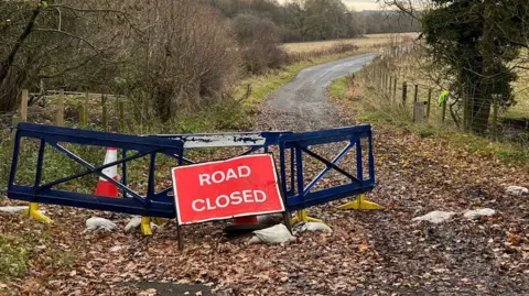 BBC Plastic road closed barriers partially block the entrance to a leaf-strewn road. One of the barriers has been moved to the side of the road.