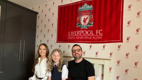 From the left a women with dark hair wearing a white shirt. In the middle, a girl with blonde hair wearing a grey uniform and on the right a man with dark hair and glasses wearing a black shirt. 

A Liverpool Football Club flag is behind them, along with a Liverpool FC wallpaper. 