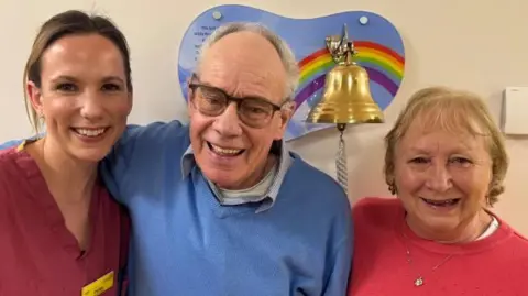 Paige Dawson, wearing a red nurse's top with her name badge on it, Bill Clarke, with short grey hair and glasses, and Alison Clarke, with short fair hair, wearing a pink top and a necklace. They are all looking at the camera and smiling. A bell on a plaque is behind them, on a wall. 