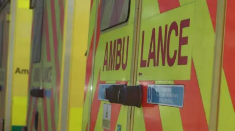 The back of a ambulance and it is orange and yellow. Over the doors the words ambulance has been written.