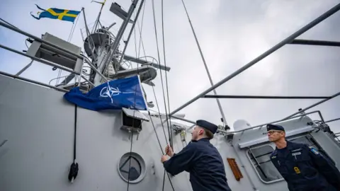 Two sailors on a military vessel. One of them is raising the NATO flag next to flag of Sweden while the other looks on. 