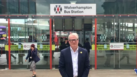 West Midlands Mayor Richard Parker, a middle-aged man with white hair and spectacles, stands in front of a set of glass doors under a sign reading "Wolverhampton Bus Station". He is wearing a blue suit jacket and a white open-necked shirt.
