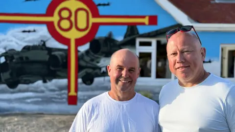 Two bald middle aged men smiling at the camera with a house in the background that has the red and yellow cross of the Guernsey flag with 80 in the middle and a large painting of an American B-17 bomber.