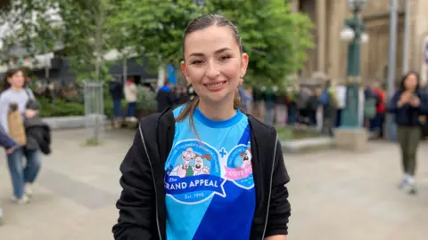 Anya Evans smiles at the camera as she stands in Bristol City Centre. She has brown hair tied back in a ponytail and is wearing a blue Wallace & Gromit Grand Appeal t-shirt underneath a black zip-up hoodie