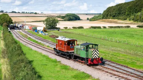 A locomotive pulls a carriage along the track, which sits beside rolling countryside.