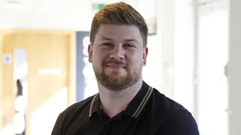 UEA Simon Rose stood in a brightly lit corridor. He has a brown spiky hair and a short beard, and is wearing a black t-shirt.