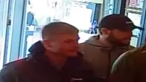 Gloucestershire Police A slightly blurry CCTV image of two men walking into a restaurant. One has a dark beard and is wearing a dark cap, while the other has his collar up and has short cropped light hair