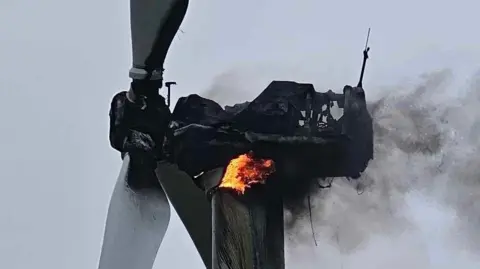 Craig Morris Close-up of the top of a wind turbine with red and orange flames at the centre. There is black smoke coming from the back of the wind turbine. The front has two white wings