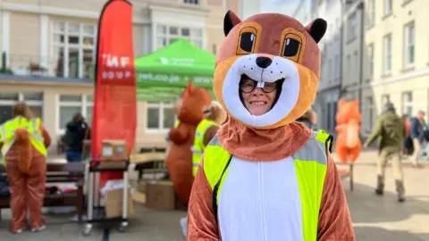 Caroline smiles at the camera wearing a full body squirrel costume and a high viz jacket. She has black framed glasses on. Behind her people are gathered by a charity stand and many people are dressed as squirrels. 