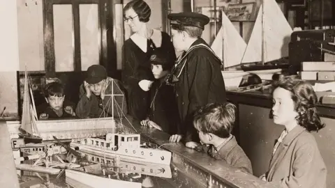 Getty Images Gelatin silver print photograph. Young boys, some wearing caps and one wearing a sailor suit, look at a toy boat display as an adult woman wearing a big collared dress and glasses keeps watch. 