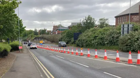 Riverside Road in Stockton is a four-lane road. There is a row of orange traffic cones in the middle, with a yellow diversion sign at the top. Cars are travelling in both directions.