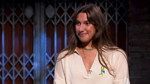 Yana Smaglo - A woman with long brown hair wearing a cream v-neck jumper and silver necklace, and a ribbon in the colours of the Ukrainian flag 