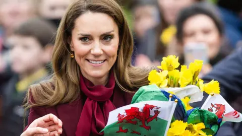 Catherine wearing burgundy carrying yellow daffodils and Welsh flags as people take pictures in the background