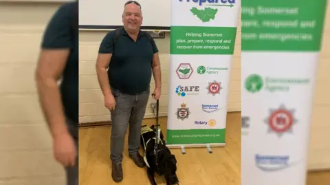 John Hardy John Hardy in a polo shirt and jeans, smiling, holding his black guide dog who is lying on the floor. He is next to a pull-up banner with Somerset emergency services on it.