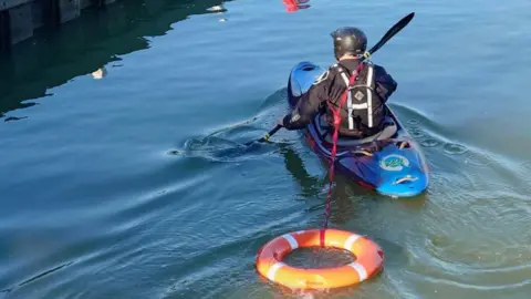 Northamptonshire Search and Rescue A man dressed in black sailing gear with a black helmet sits in a blue single-person kayak rowing in a river with an oar. A piece of red rope is stretched between the craft and an orange buoy which is being pulled along to bank.