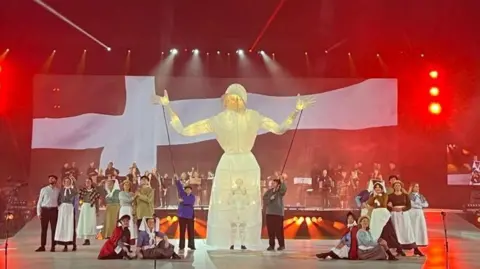 A large stage with red beaming lights on the left and right side of the image. There is a large black and white Cornish flag in the background, and in front of it is a big white puppet, and a crowd of performers on the stage in old-fashioned clothing. 