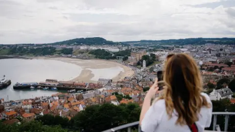 The image shows an elevated view over Scarborough, with a sweeping bay, sandy beach, harbour, and clusters of rooftops stretching across the landscape. In the foreground, a woman with long hair, seen from behind, stands at a railing, holding up a phone to take a photo of the scenery.