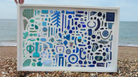 University of Brighton Various shade of blue sea plastic arranged and stuck to a white framed background. A hand is holding the top left of the frame to keep it upright. It is on the pebbles of a beach. The sea is in the background