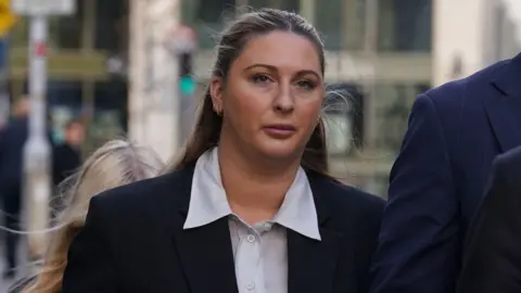 PA Media Ms Hand walking through the court gates, she has long blonde hair and is wearing makeup. She is wearing a black suit jacket with a blue shirt.