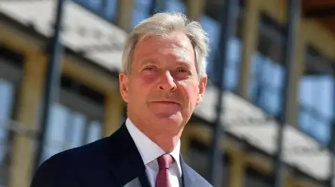 LDRS A man with short grey hair wearing a black suit and a red tie is standing in front of a building
