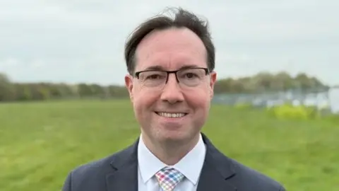 Qays Najm/BBC Mark Flynn wears glasses and stands in a field. He wears a navy suit jacket, light-coloured check-patterned tie and a pale blue shirt. He is smiling at the camera.