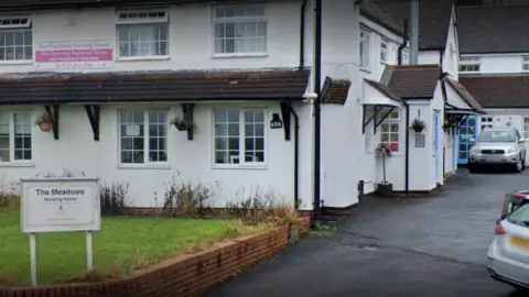 The outside of a white rendered building with brown roof tiles. A car park is visible to the right hand side. On the left is a small patch of lawn with a sign that says The Meadows Nursing Home