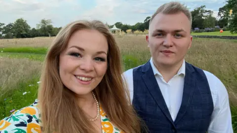 EMILY HANDSTOCK Woman with long hair in bright floral dress, with her husband who has short blonde hair and is wearing a waistcoat and shirt. They smile at the camera on a sunny day, standing in a field.