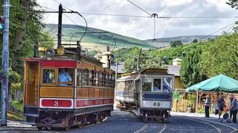 Manx Scenes Two electric trams sit side by side on tracks with a canopy on the right and people waiting. In the background there are trees and rolling hills.