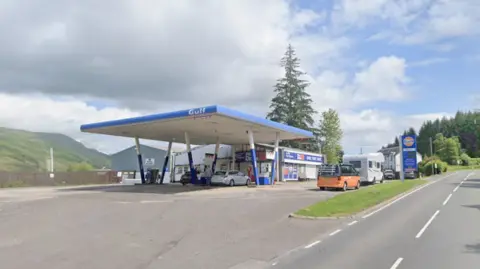 Google Maps Gulf petrol station with a blue canopy over multiple fuel pumps beside a roadside, with cars parked at the forecourt, a shop building behind, green hills in the background, and a partly cloudy sky.
