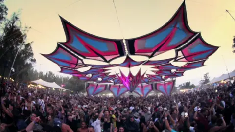 It is still a footage of the Nova Festival before the attack, with a large crowd dance under a star -shaped umbrella in narcotic and pink tones. Some lights are lit and the sky has a touch of early morning glow