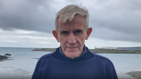 Peter Watson is looking serious as he stands in St Sampson with the sea behind him on a grey day. He has short grey hair and is wearing a navy Guernsey sweater.