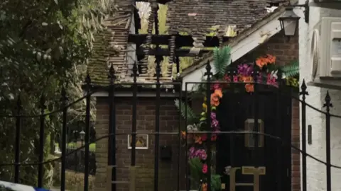 A restaurant with a timber roof and flowers on the front. The roof has been damaged by fire and is caged in, with trees visible through the gaps.