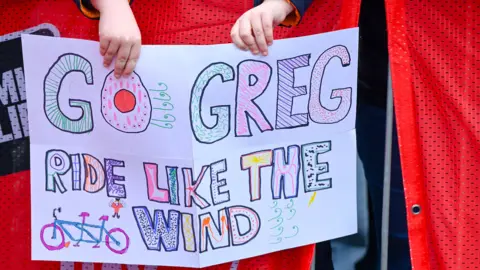 A child's hands holding a hand-drawn sign that reads 'Go Greg, ride like the wind'.