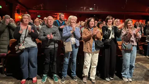 A crowd of people of different ages and genders stand and applaud in the auditorium of a cinema. 