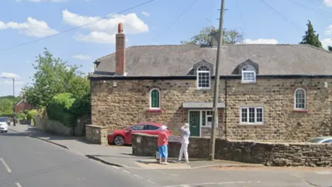 Two people stand at the corner of two roads on a sunny day. There are large stone-coloured houses and a red car parked on the drive.